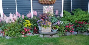 Un jardín con diversas plantas y flores frente a una casa con revestimiento oscuro y detalles blancos.