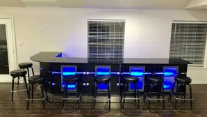 Uma área de bar com bancada preta, iluminação azul de LED e seis banquetas pretas.