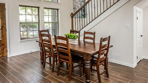 Uma sala de jantar com mesa e cadeiras de madeira, um vaso de planta e uma escada ao fundo.