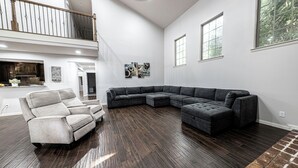 Uma sala de estar ampla com um sofá seccional, uma poltrona cinza e piso de madeira.