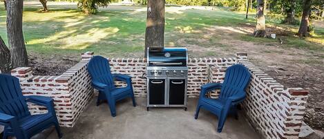 An outdoor patio with a grill, two blue chairs, and a brick wall.