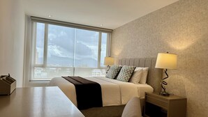 A modern hotel room with a large window, a bed with two pillows, a bedside table with a lamp, and a wooden nightstand.