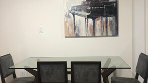 A dining area with a glass-top table and four chairs, a painting of a piano on the wall, and a white ceiling. - Relaxed and Spacious 2 BR Apartment with a Patio (Oakland)
