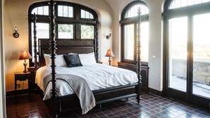 Una cama con dosel, un cabecero de madera, dos mesitas de noche con lámparas y ventanas arqueadas grandes.