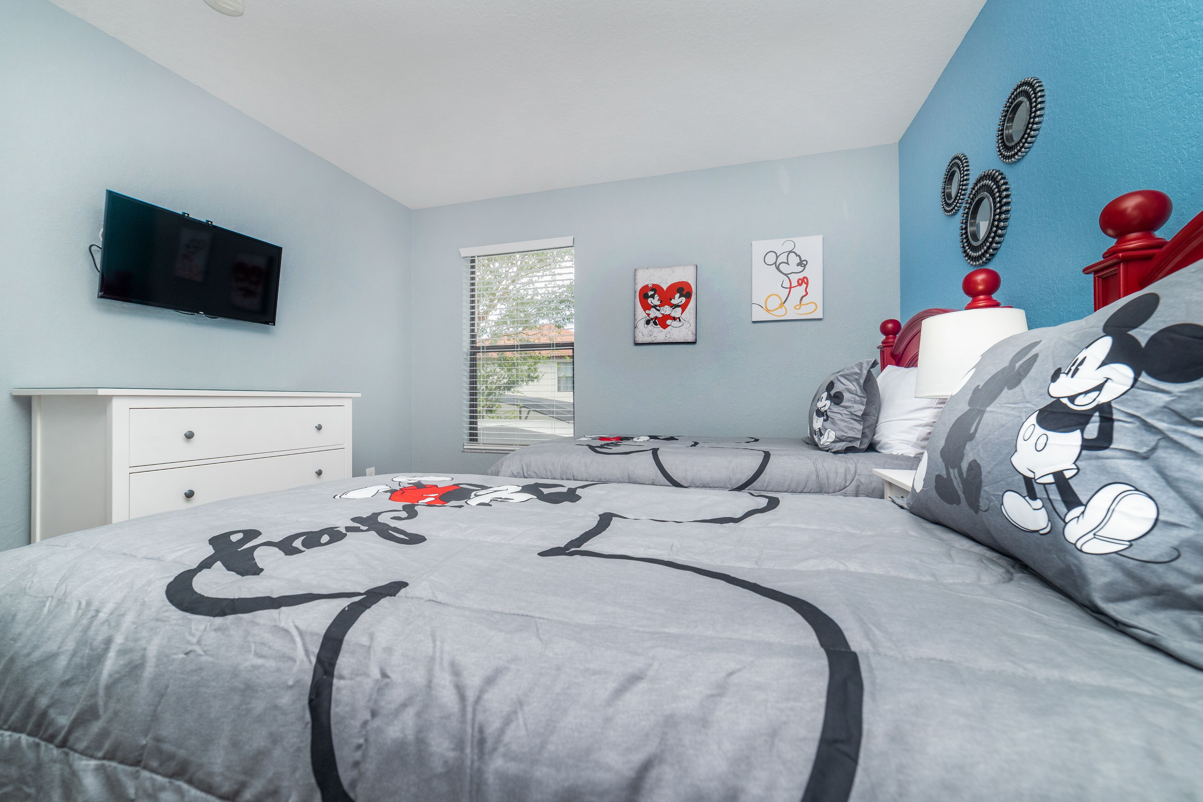 A bedroom with a bed featuring Mickey Mouse designs, a flat-screen TV mounted on the wall, and a window with blinds.
