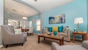 Una sala de estar con un sillón gris, un sofá color beige con cojines de colores, una mesa de centro y una lámpara. Un cuadro cuelga en la pared, y se ve un reloj.