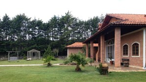 Une maison avec un toit en tuiles, des murs en briques et un porche couvert. On y trouve une pelouse avec quelques palmiers et un kiosque en arrière-plan.