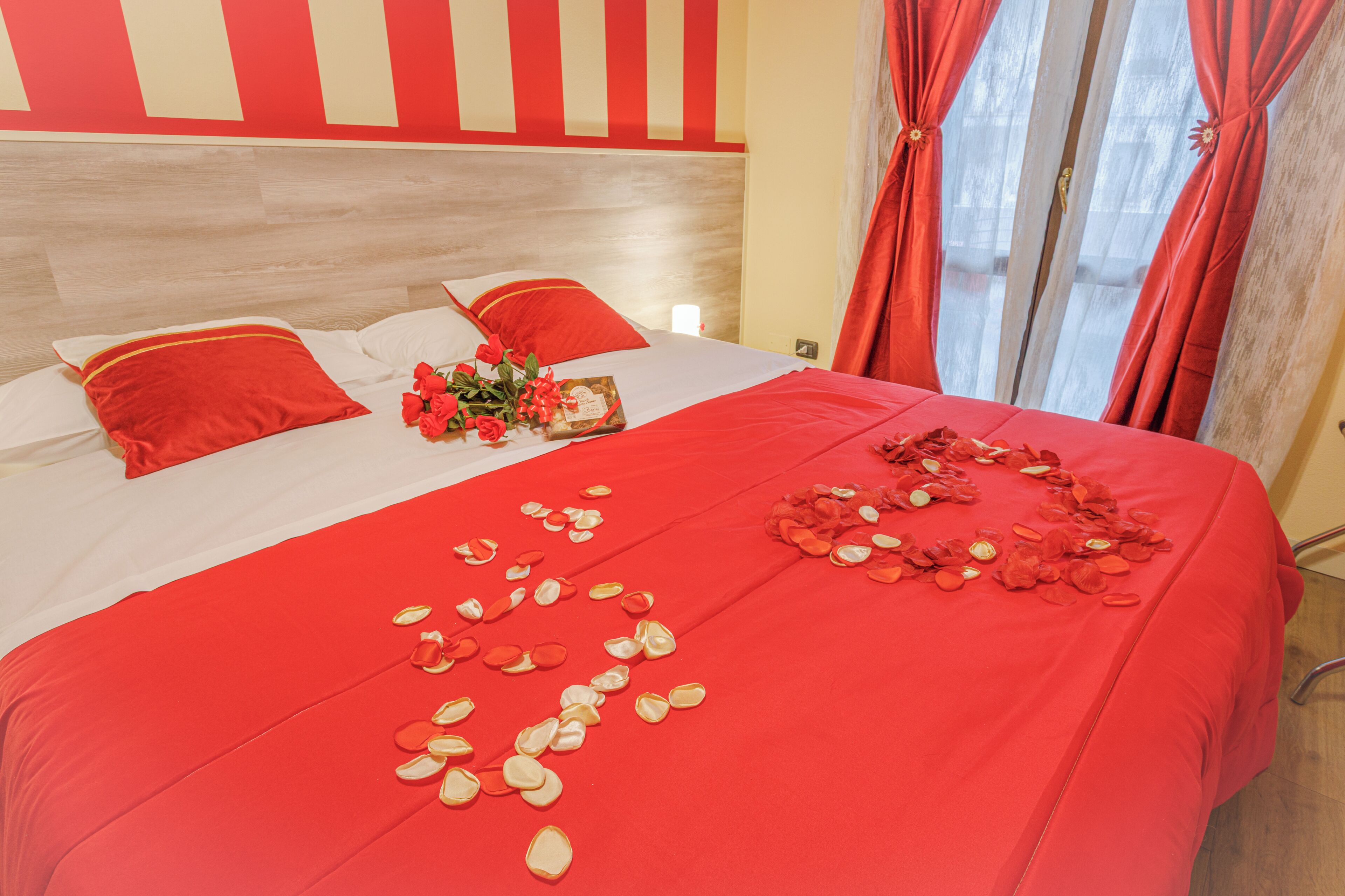 A bed with red bedding, a bouquet of flowers, and scattered rose petals.