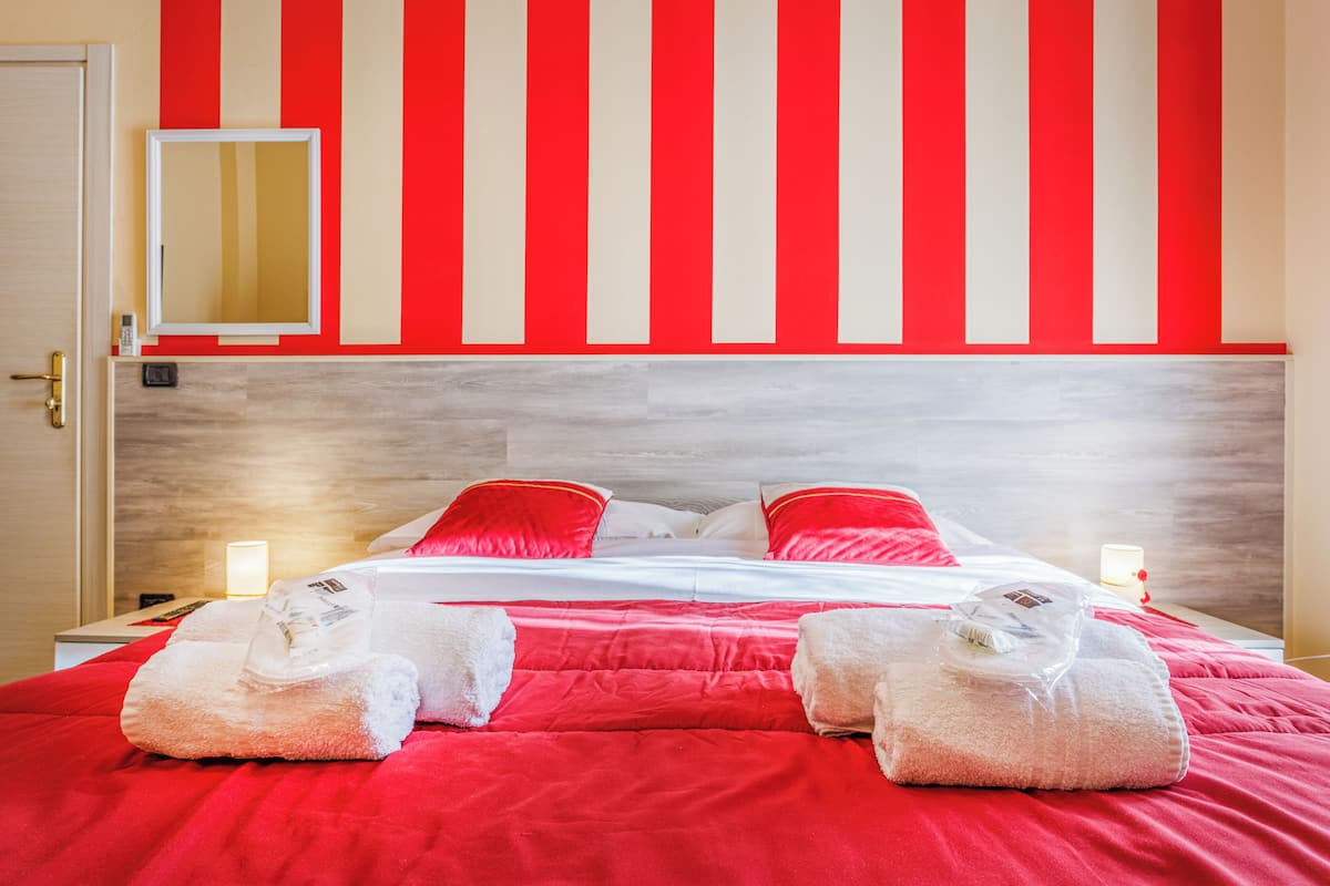 Una habitación de hotel con cama, almohadas, una mesita de noche, un espejo y una pared con rayas verticales.