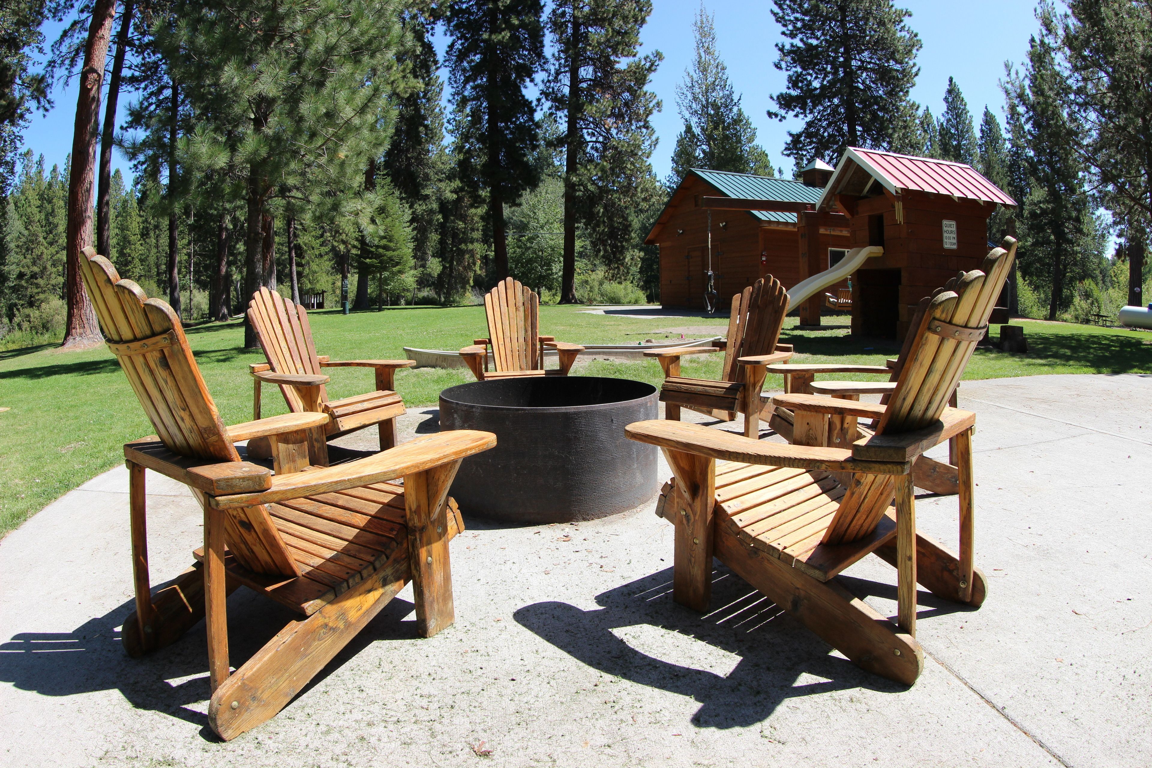 A fire pit surrounded by wooden Adirondack chairs in a forested area with a cabin in the background.