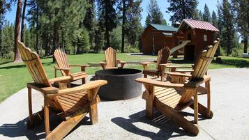 A fire pit surrounded by wooden Adirondack chairs in a forested area with a cabin in the background.