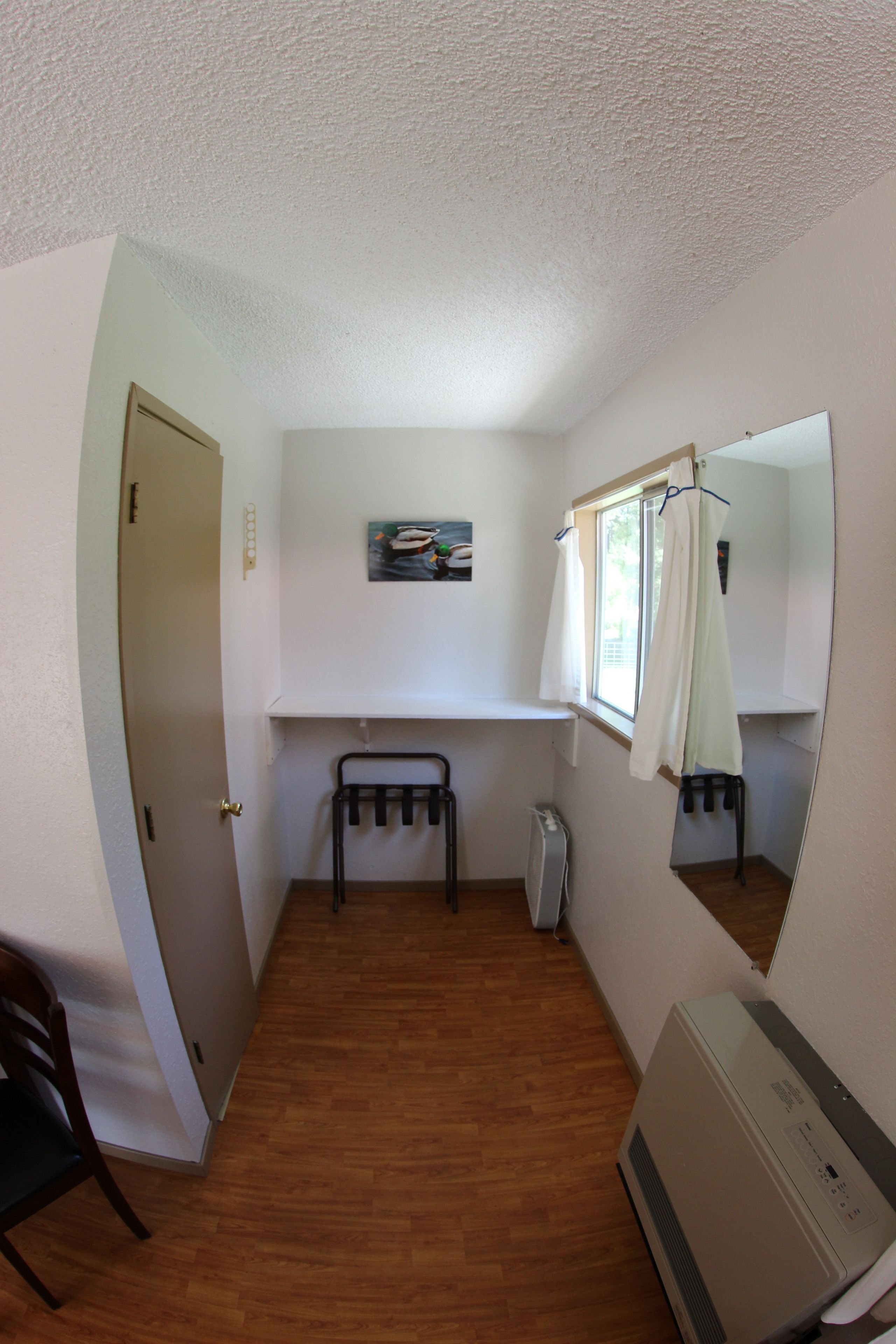 Una habitación pequeña y funcional con suelo de madera, un radiador blanco, una mesita, una silla, un espejo y una puerta.