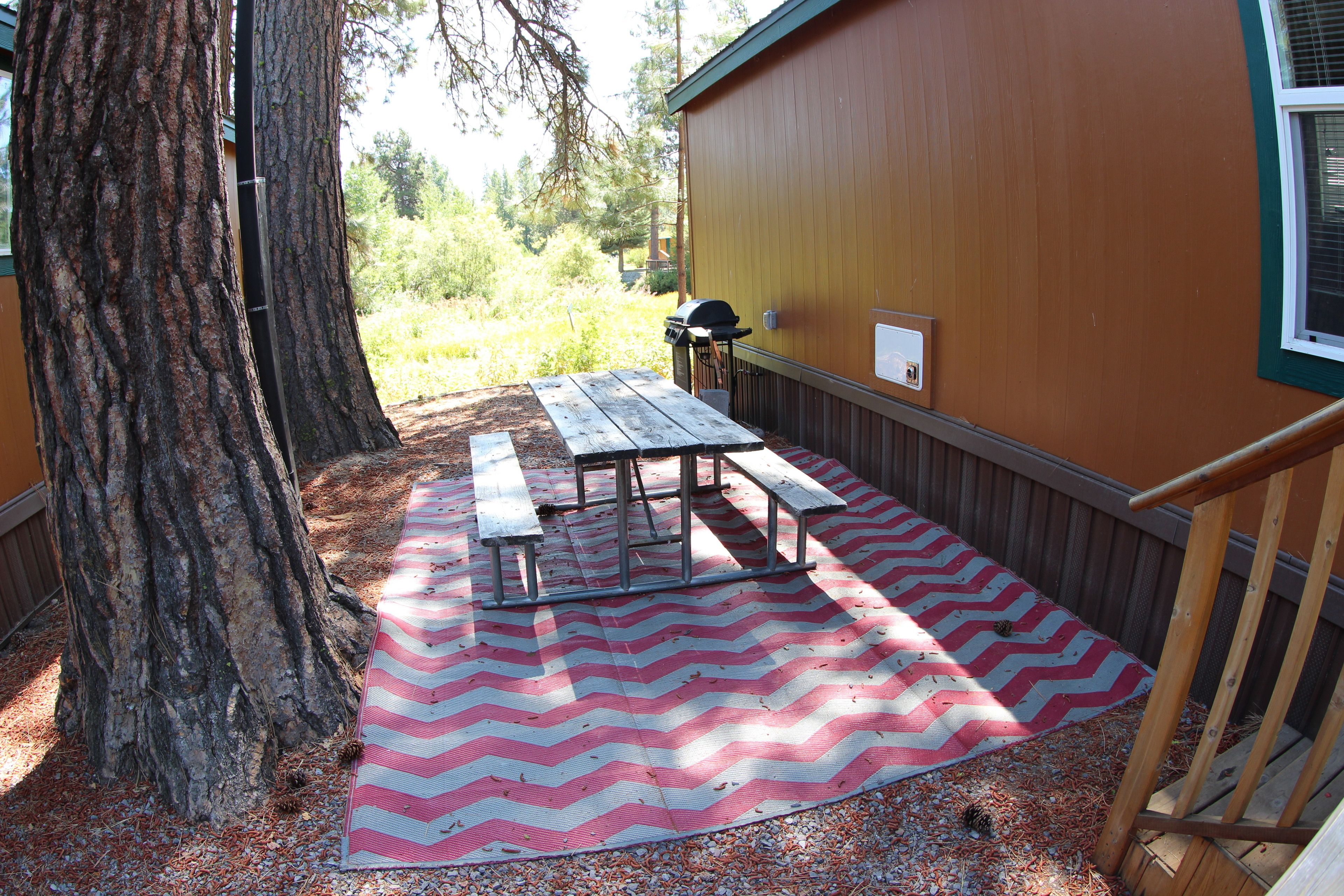 A picnic table set up on a chevron-patterned mat, a wooden bench, and a barbecue grill against a building with a window.