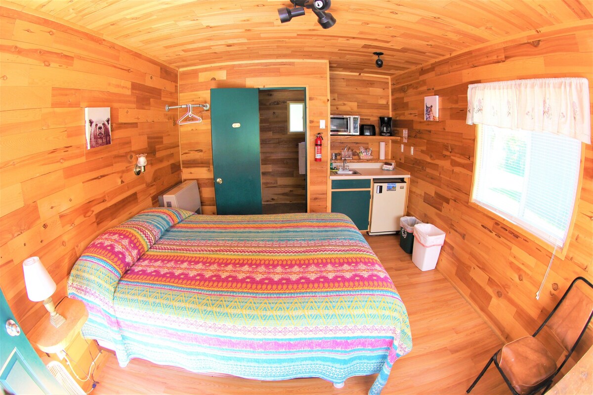 A cozy wooden cabin room with a bed, a small kitchenette, a bathroom, and a window with curtains.