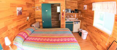 Una habitación acogedora tipo cabaña de madera, con una cama, una kitchenette pequeña, un baño y una ventana con cortinas.