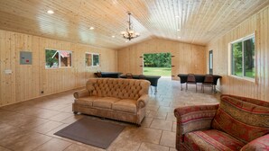 A spacious room with wooden walls and ceiling, a leather sofa, a patterned sofa, a dining table with chairs, and a large window offering a view of the outdoors. - Crater Lake Resort (Chiloquin)