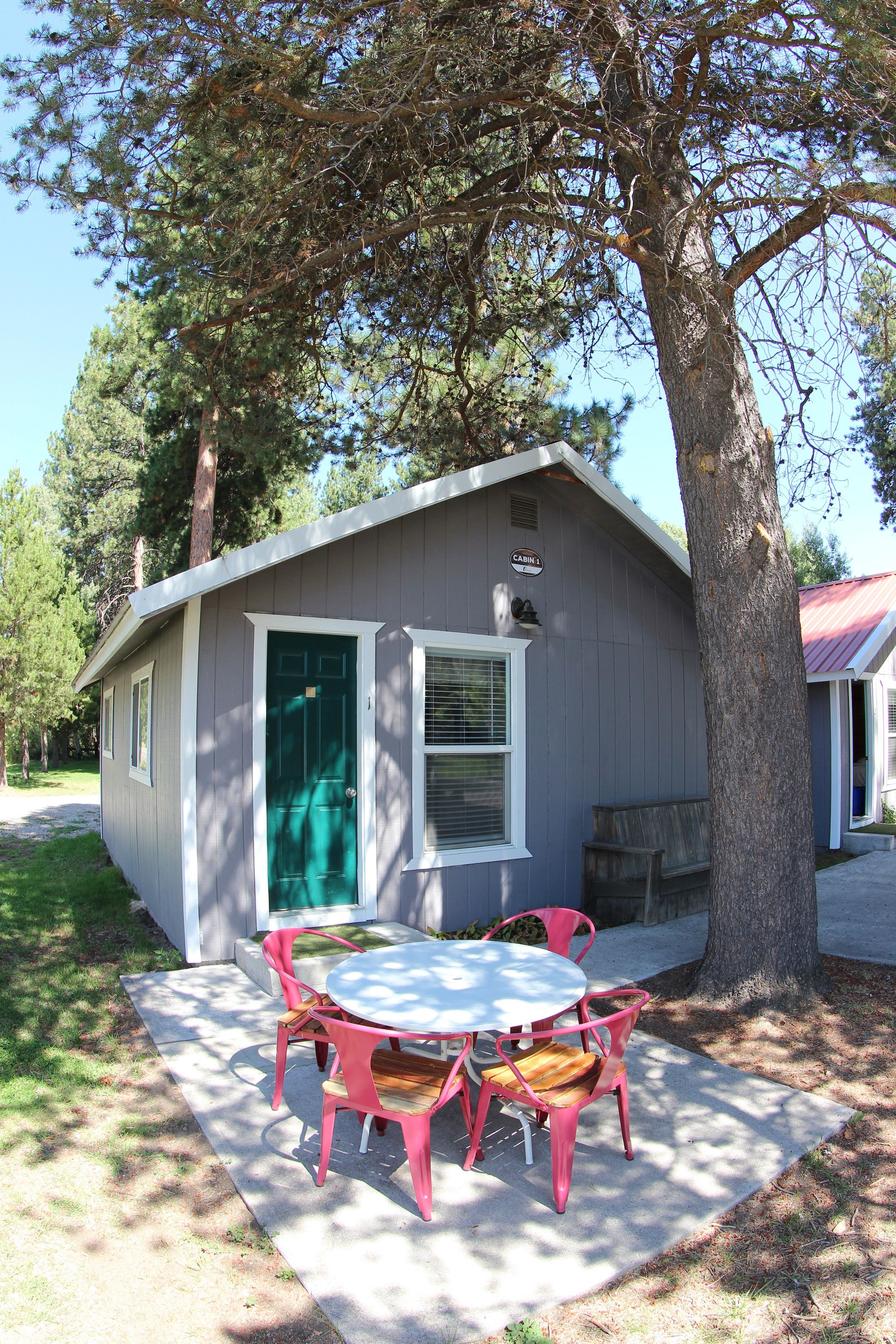 Une petite cabane avec une porte verte, une table ronde entourée de chaises roses et un arbre devant.