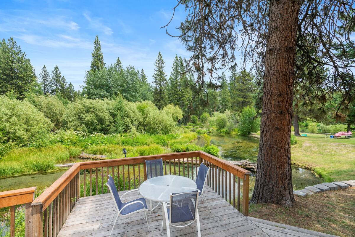 A wooden deck with a glass table and chairs overlooking a pond and forest.