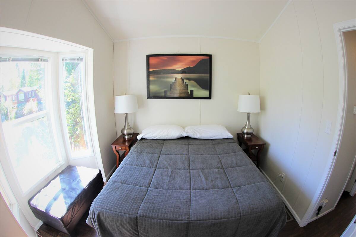 A bedroom with a large bed, two bedside lamps, a window with a view of greenery, and a painting of a landscape on the wall.
