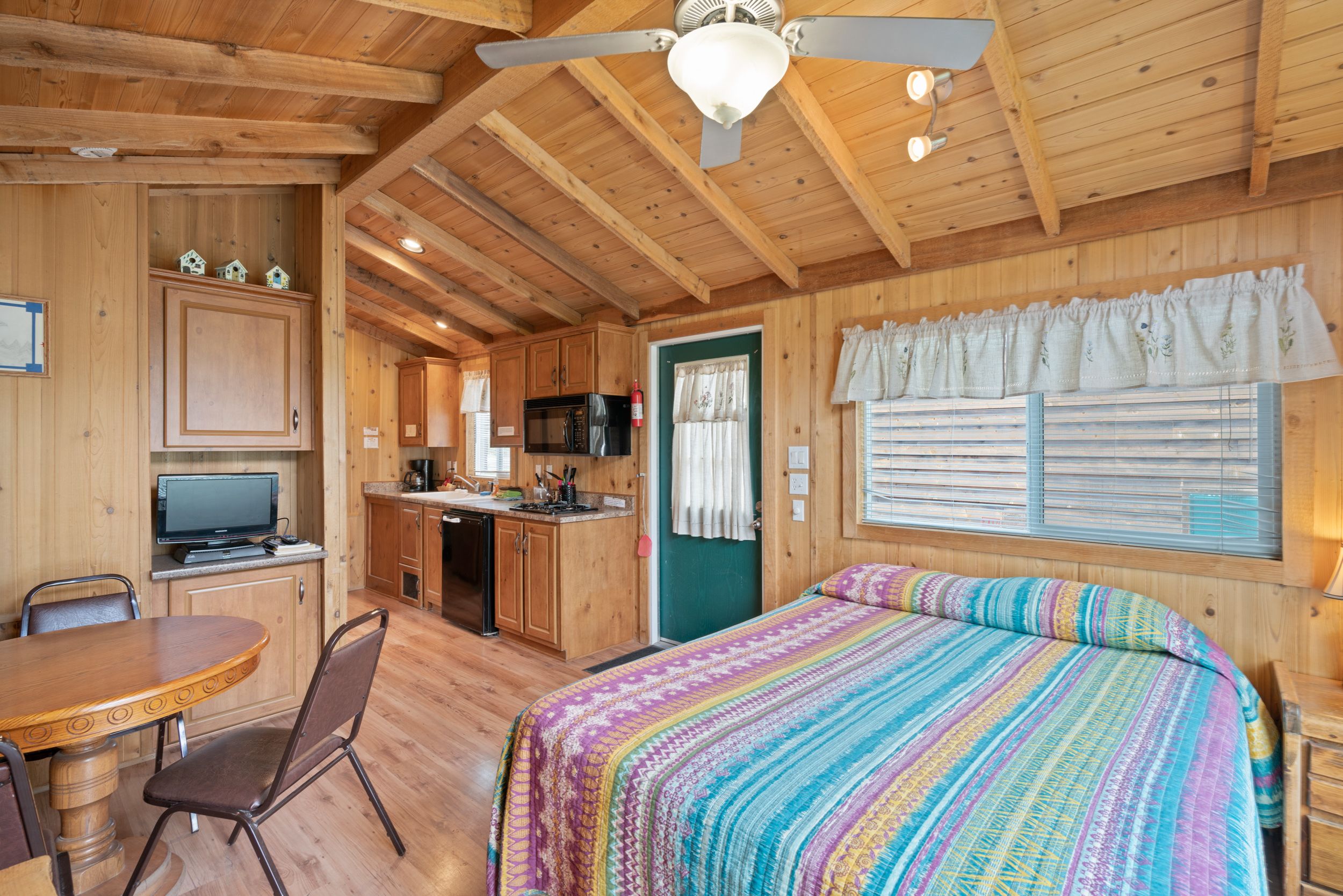 Une cabane en bois comprenant un coin repas, une cuisine et un grand lit recouvert d’une couverture colorée.