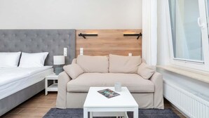 A modern hotel room with a grey tufted headboard bed, a beige sofa, a small white table with a magazine, and a window with blinds. - Cozy Studio 150 m from the Beach | Balcony and Parking (Międzyzdroje)
