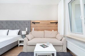 Una habitación de hotel moderna con una cama y cabecera gris capitonada, un sofá beige, una pequeña mesa blanca con una revista y una ventana con persianas.
