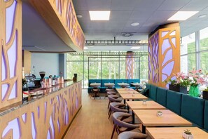 Un restaurante moderno con mesas de madera, asientos acolchados verdes, ventanas amplias y paneles decorativos en las paredes.