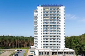 Un hotel de varios pisos con balcones, un estacionamiento y un fondo arbolado.