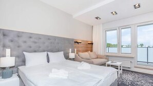 A modern hotel room with a large bed, a grey tufted headboard, a sofa, and a view of the city through large windows. - Cozy Studio 150 m from the Beach | Balcony and Parking (Międzyzdroje)