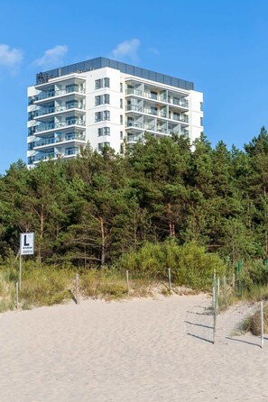 Ein mehrgeschossiges Gebäude mit Balkonen, umgeben von dichten Kiefern und einem Sandstrand.