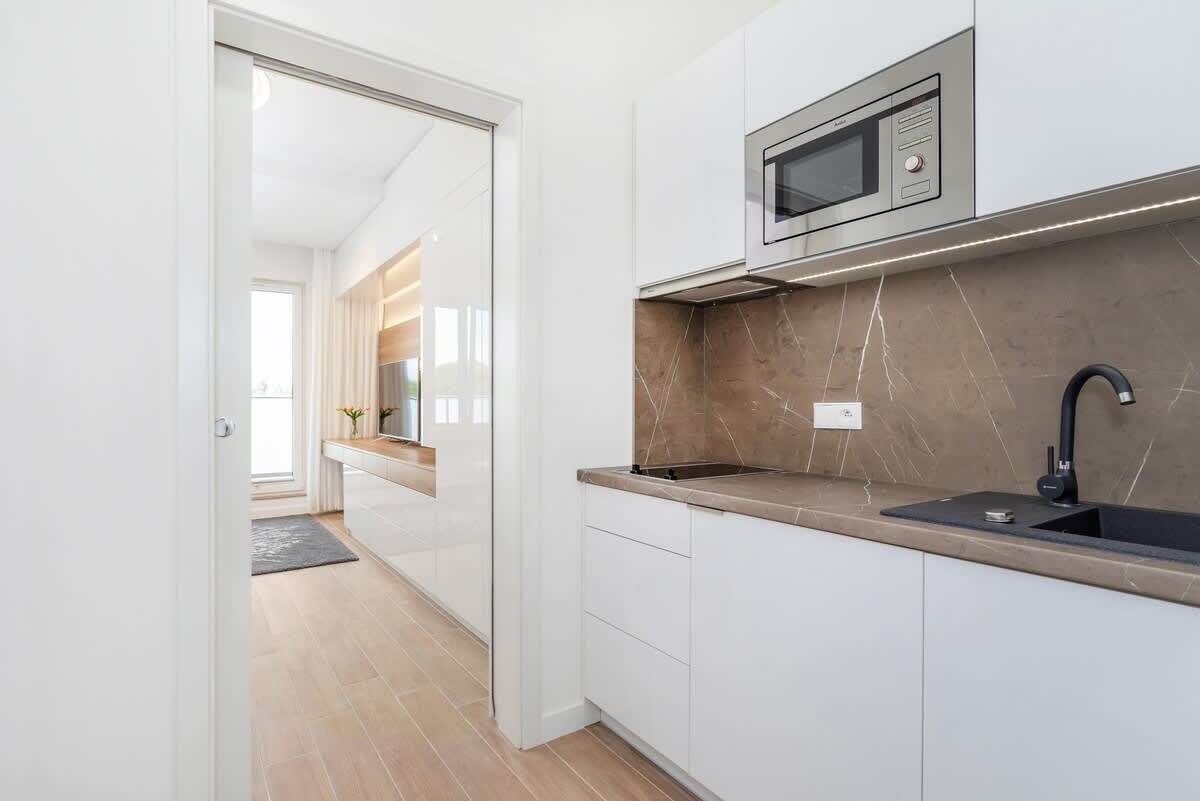 Una cocina moderna con armarios blancos, protector contra salpicaduras de mármol, microondas empotrado y un lavamanos negro.