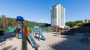 A playground with slides and climbing structures, a sandy area, and a tall building in the background. - Cozy Studio 150 m from the Beach | Balcony and Parking (Międzyzdroje)