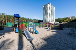 Un parque infantil con toboganes y estructuras para escalar, una zona de arena y un edificio alto al fondo.