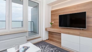 Una sala de estar moderna con un televisor de pantalla plana grande montado en una pared de madera, una mesa de centro blanca con una revista, y vistas al balcón a través de ventanas amplias.