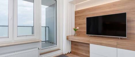 A modern living room with a large flat-screen TV mounted on a wooden wall, a white coffee table with a magazine, and a view of the balcony through large windows.