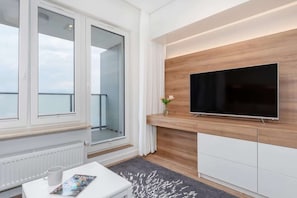Una sala de estar moderna con un televisor de pantalla plana grande montado en una pared de madera, una mesa de centro blanca con una revista, y vistas al balcón a través de ventanas amplias.