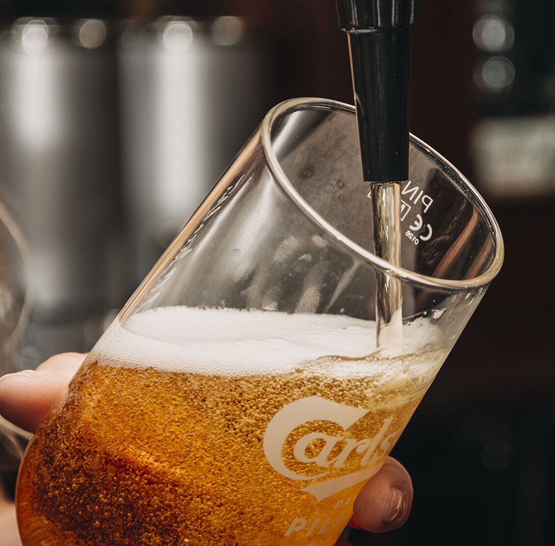 Uma mão segurando um copo com um tubo de chopp, cheio de cerveja e espuma, e um logotipo no copo.