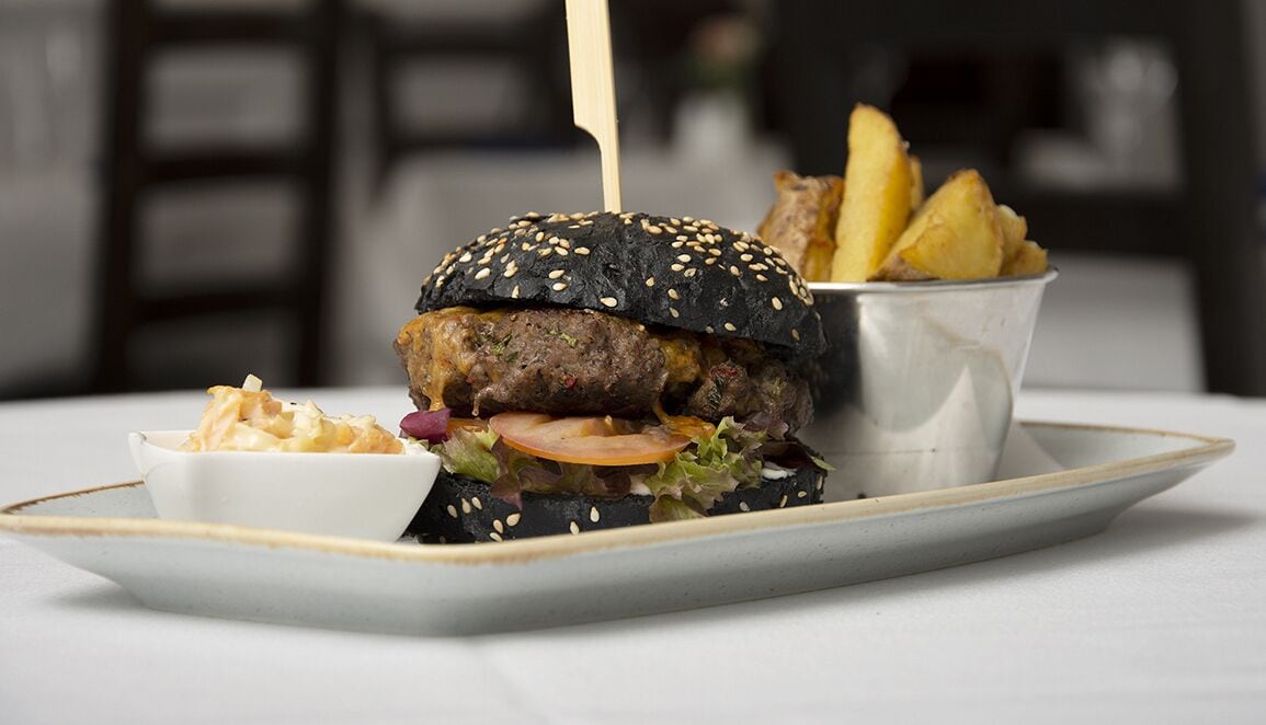 Un hamburger servi dans un pain aux graines de sésame, garni de laitue, de tomate et accompagné d’un cure-dent, le tout présenté sur une assiette avec des frites et un petit ramequin de sauce.