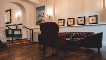 A room with a wooden floor, a red armchair, a brown leather sofa, a small table with a bottle, and framed pictures on the wall.