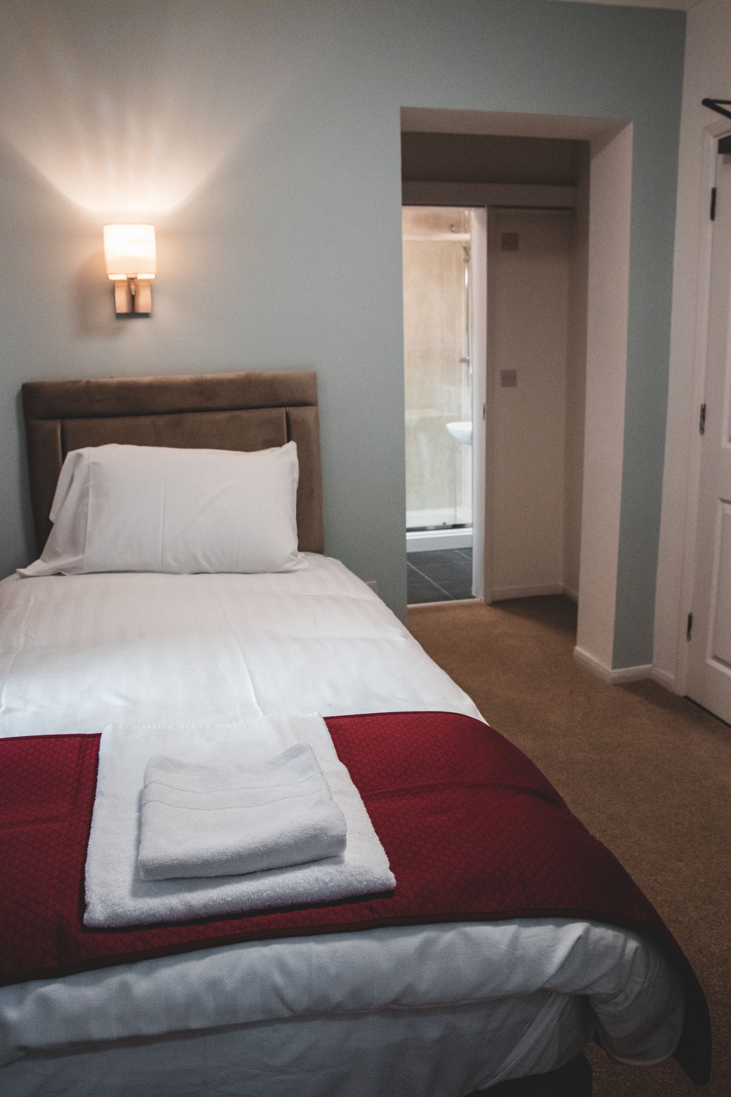 Una cama bien tendida con sábanas blancas y un edredón rojo, una lámpara de pared y una puerta que da a un baño.