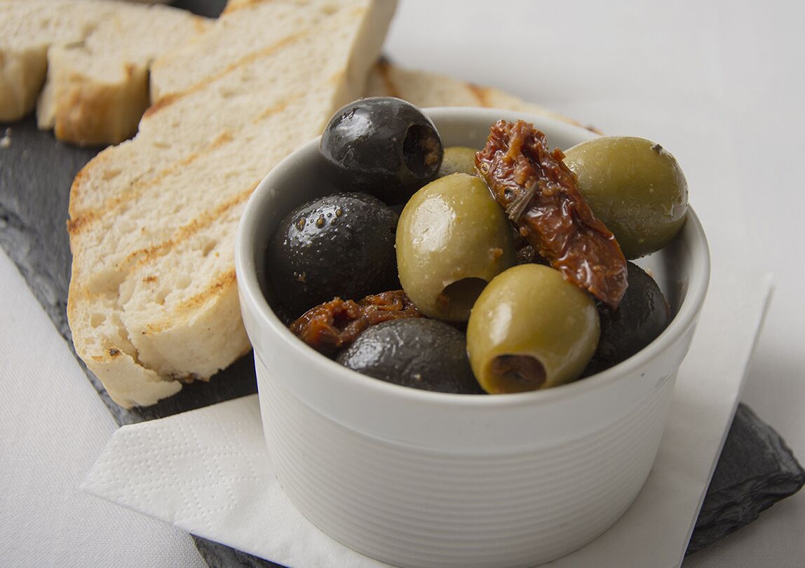 Een schaal olijven en zongedroogde tomaten op een arduinen bord met sneetjes brood.