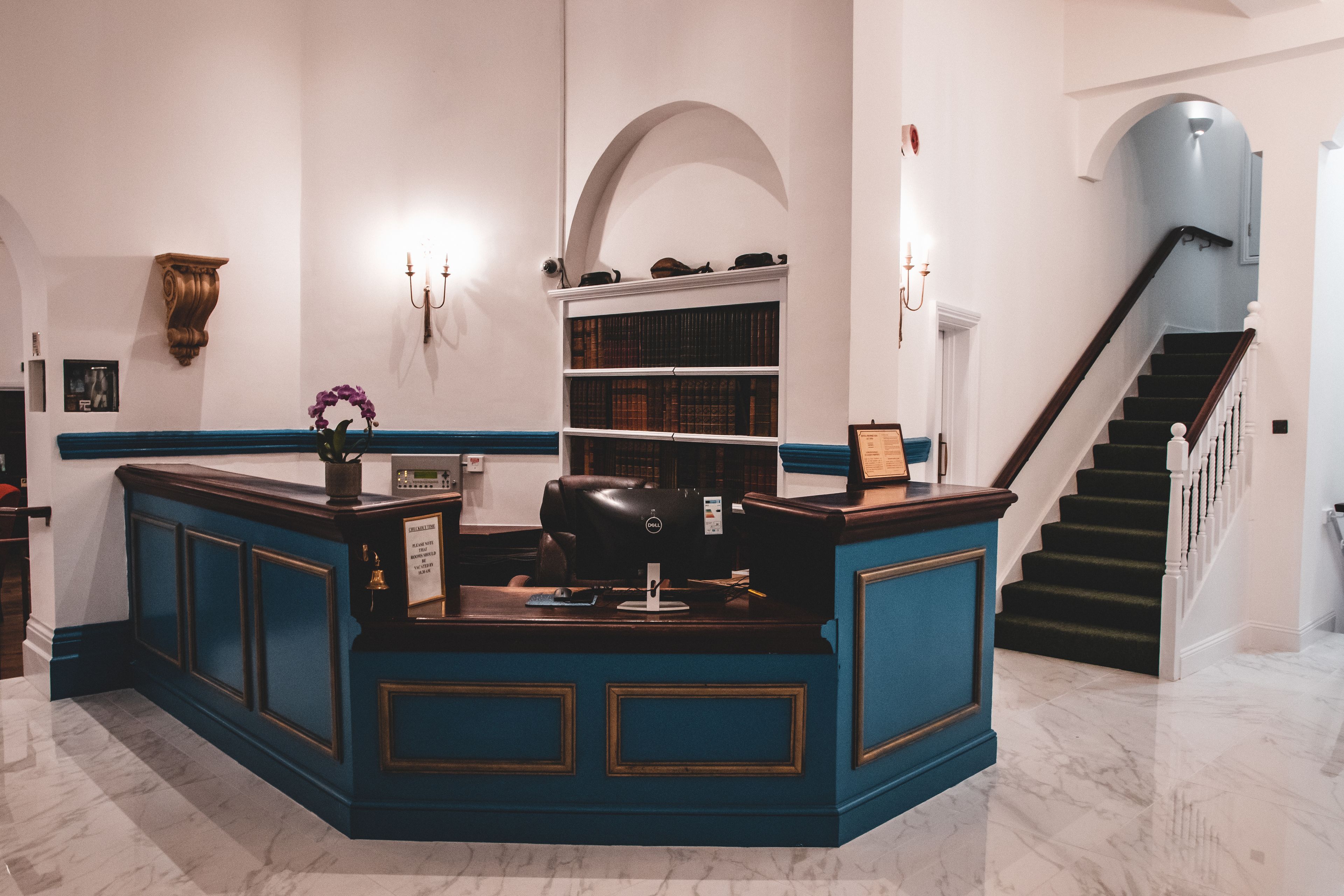 Een receptiebalie met blauw en goud decor, een boekenplank vol boeken, een trap en een kroonluchter.