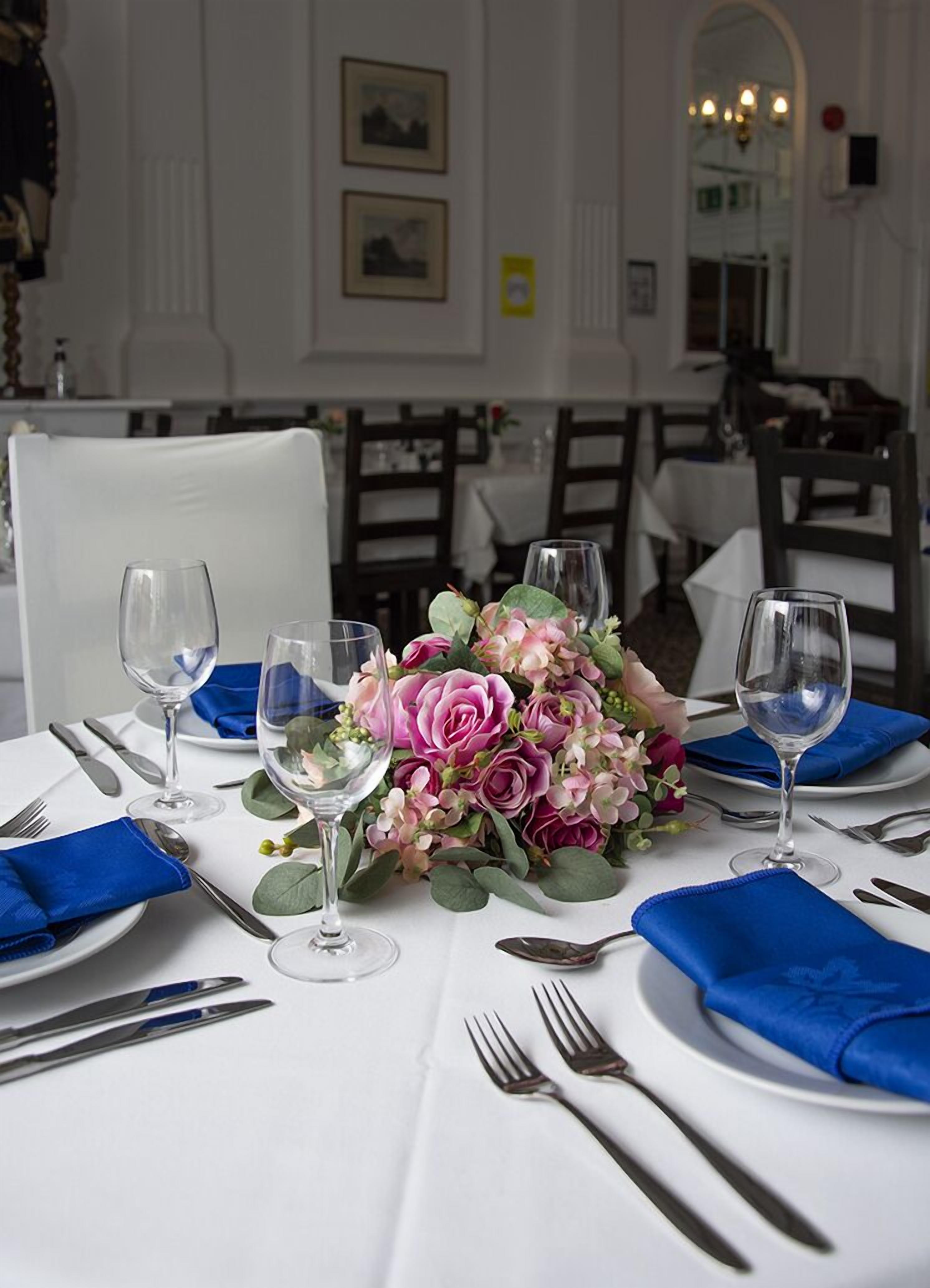 A dining table set with blue napkins, silverware, and wine glasses, with a floral centerpiece.