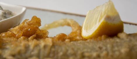 Primer plano de un pescado frito con una rodaja de limón encima, acompañado de un bol de salsa.