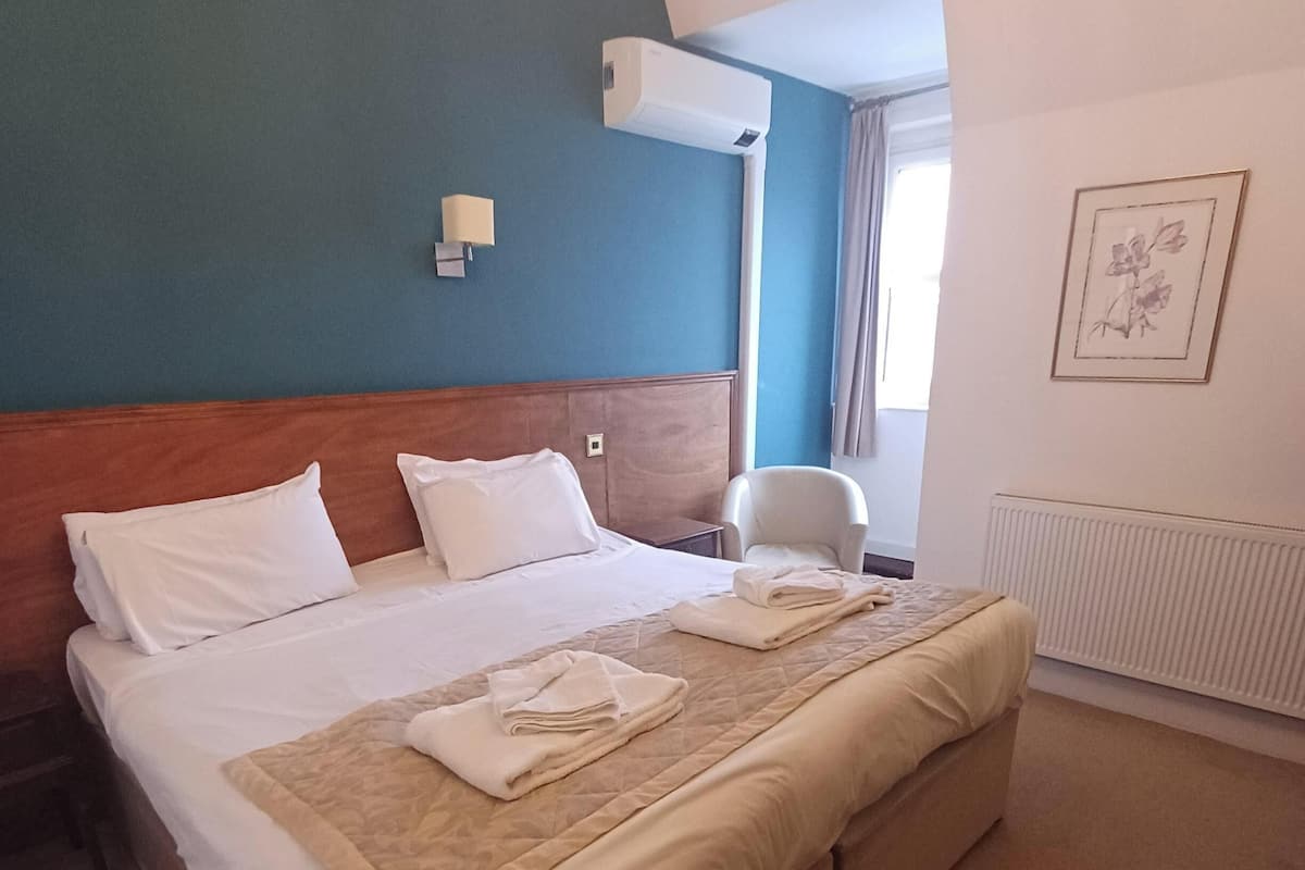 Hotellihuone, jossa on suuri sänky, puinen pääty, seinälle asennettu ilmastointilaite, pieni pöytä, tuoli ja seinällä oleva kehystetty kuva.