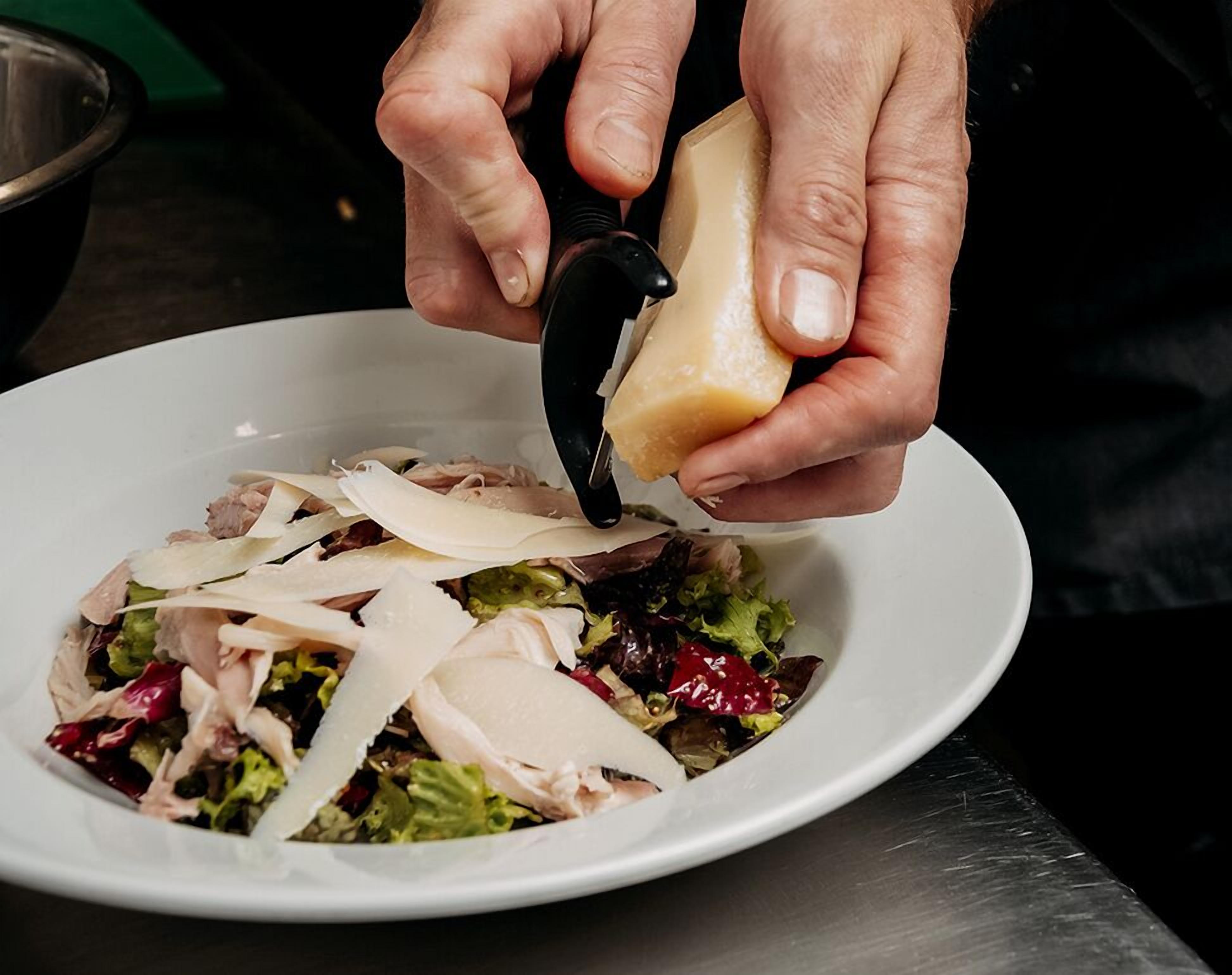 Eine Person schneidet ein großes Stück Käse über einen Salat in einer weißen Schüssel.