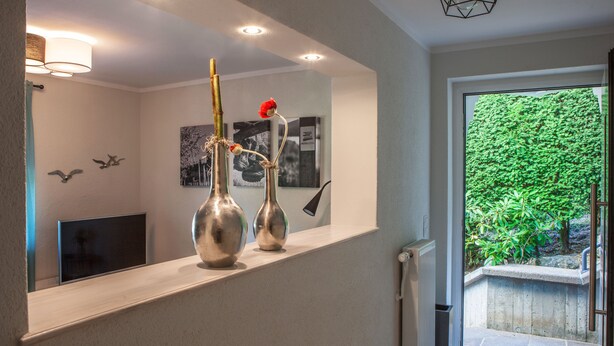 Un intérieur moderne avec une console en bois sur laquelle sont disposés deux vases métalliques, une photographie en noir et blanc encadrée, et une vue sur un jardin verdoyant à travers une porte vitrée.