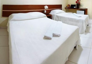 Duas camas com lençóis e toalhas brancas, um cabeceira de madeira, uma mesa de cabeceira com abajur e uma pequena escrivaninha com computador e objetos decorativos.