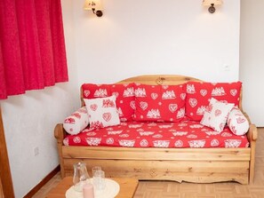Um sofá de madeira com almofadas estampadas em vermelho e branco, uma mesa de madeira com copos e uma vela, e uma cortina vermelha numa parede branca.