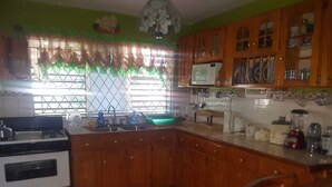 A kitchen with wooden cabinets, a stove, a sink, and a window with curtains. - Victoria's Place (Portmore)